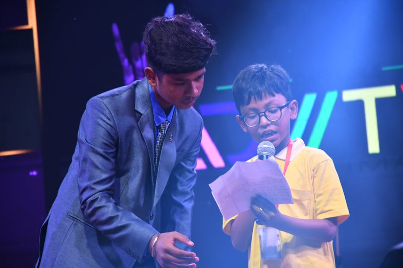 A young student reading a speech on stage, helped by a senior student